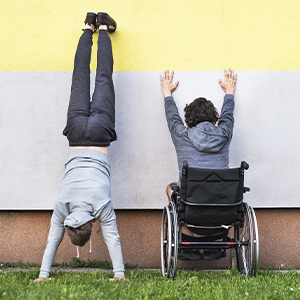 Young man in wheelchair facing a wall with his hands up it and another young man facing wall doing a handstand. they are side by side.