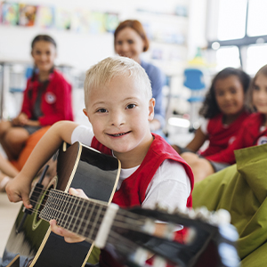 Young boy Down’s syndrome playing guitar with other children and teacher in the background